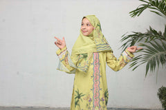 Young girl in a green traditional outfit with a hijab, pointing to the side against a white wall with a plant.