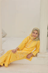 Woman in a yellow traditional outfit with embroidery sitting on a white bed.
