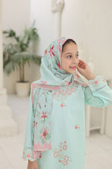 Young girl wearing a light blue floral hijab and dress in a bright indoor setting.