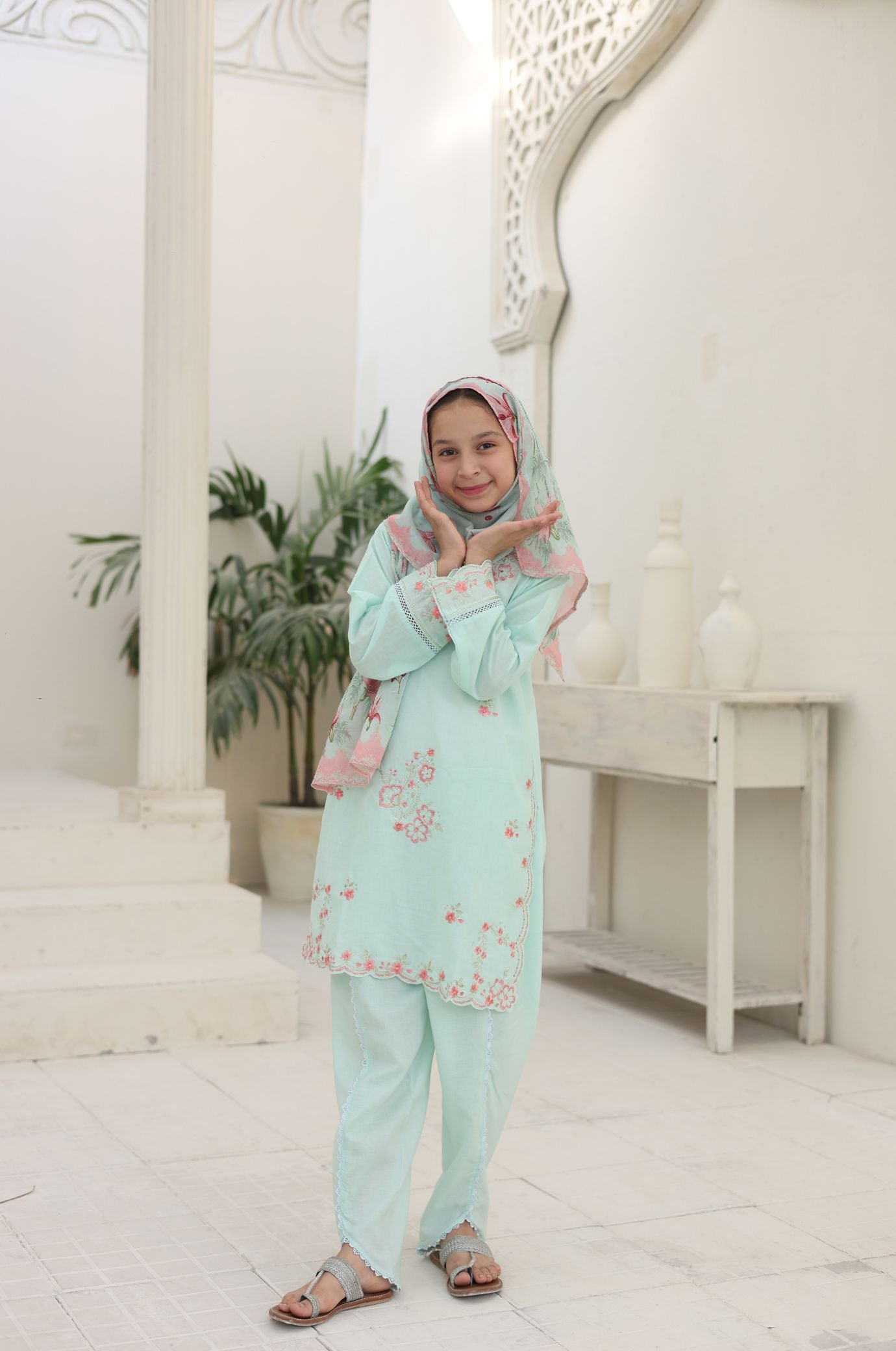 Woman in a light green traditional outfit with a pink hijab standing indoors.