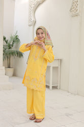 Young girl in a yellow traditional outfit with a headscarf, standing in a white interior setting.
