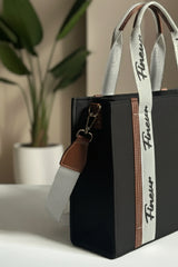 Black handbag with a white strap featuring 'fineur' branding, placed on a surface with a plant in the background.