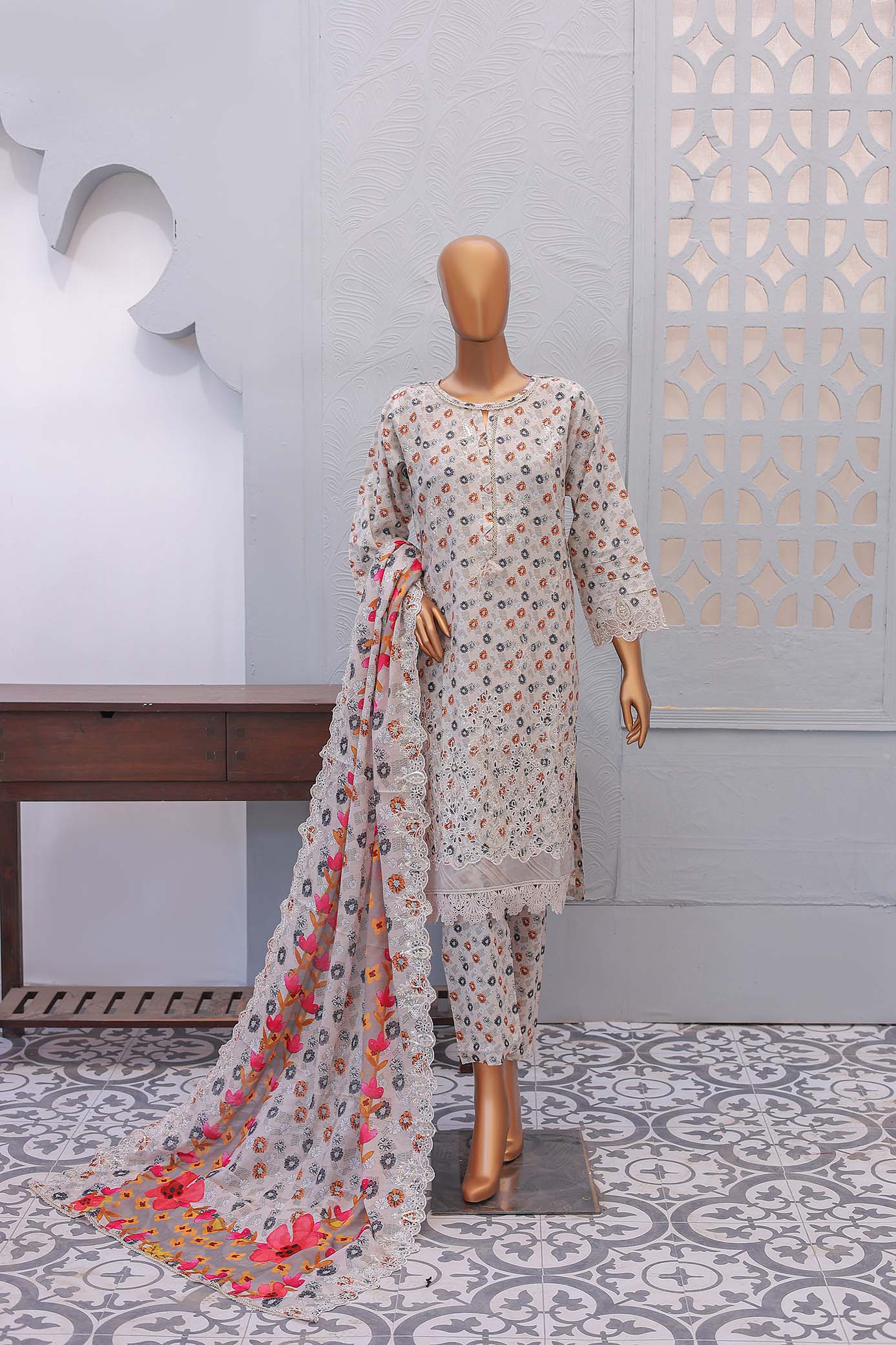 A three-piece beige festive embroidered outfit consisting of a shirt with front and sleeve embroidery, laser and cutwork embellishments, trousers, and a dupatta with printed cutwork, displayed on a mannequin against an indoor backdrop.