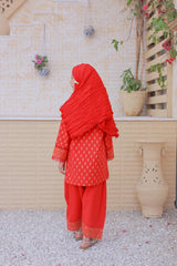 Person wearing a red traditional outfit standing in front of a beige wall with plants.