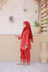 Person wearing a red traditional outfit with a matching headscarf, standing against a light-colored wall with plants.