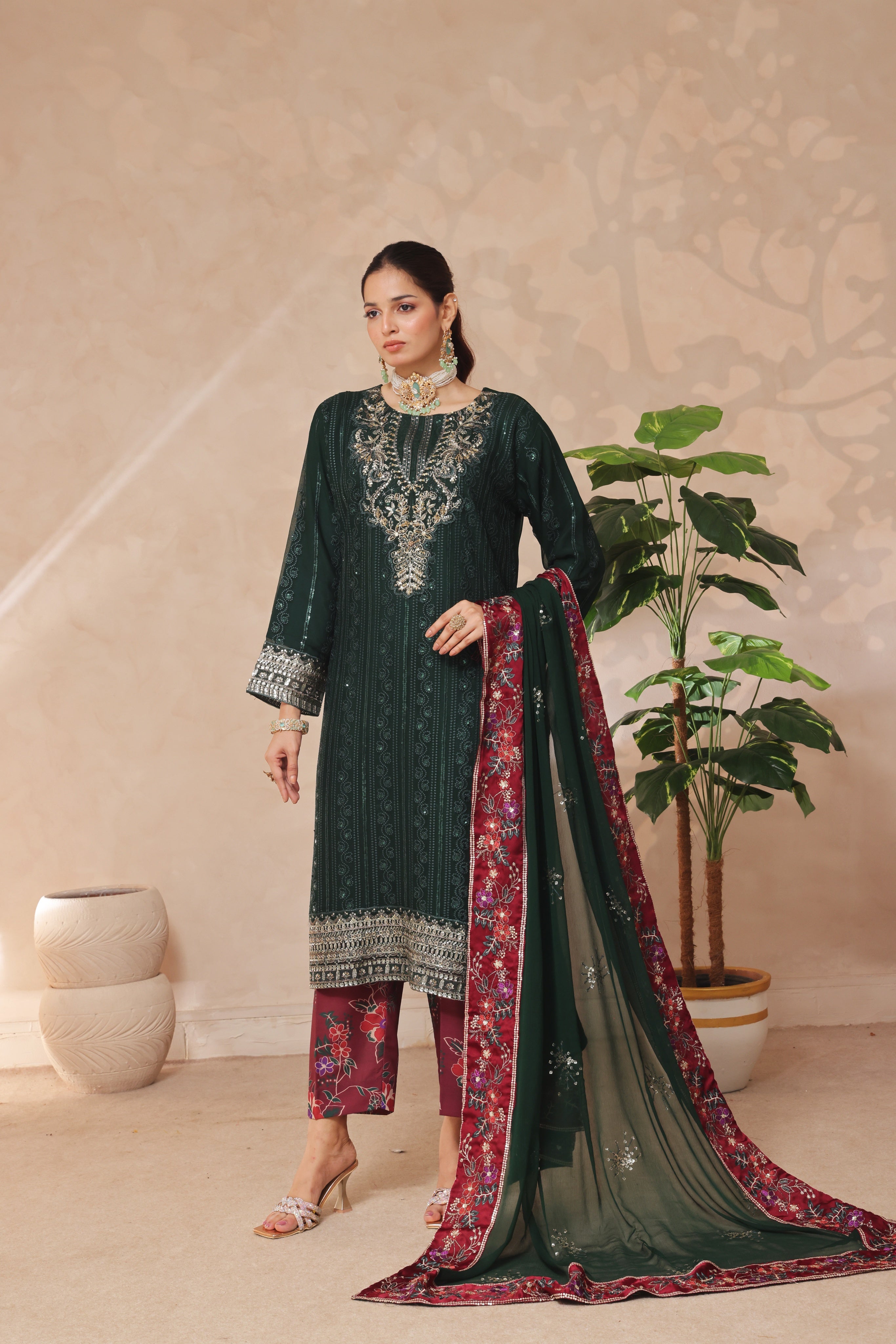 Woman in a dark green embroidered outfit with a red and gold dupatta in a room with a plant.