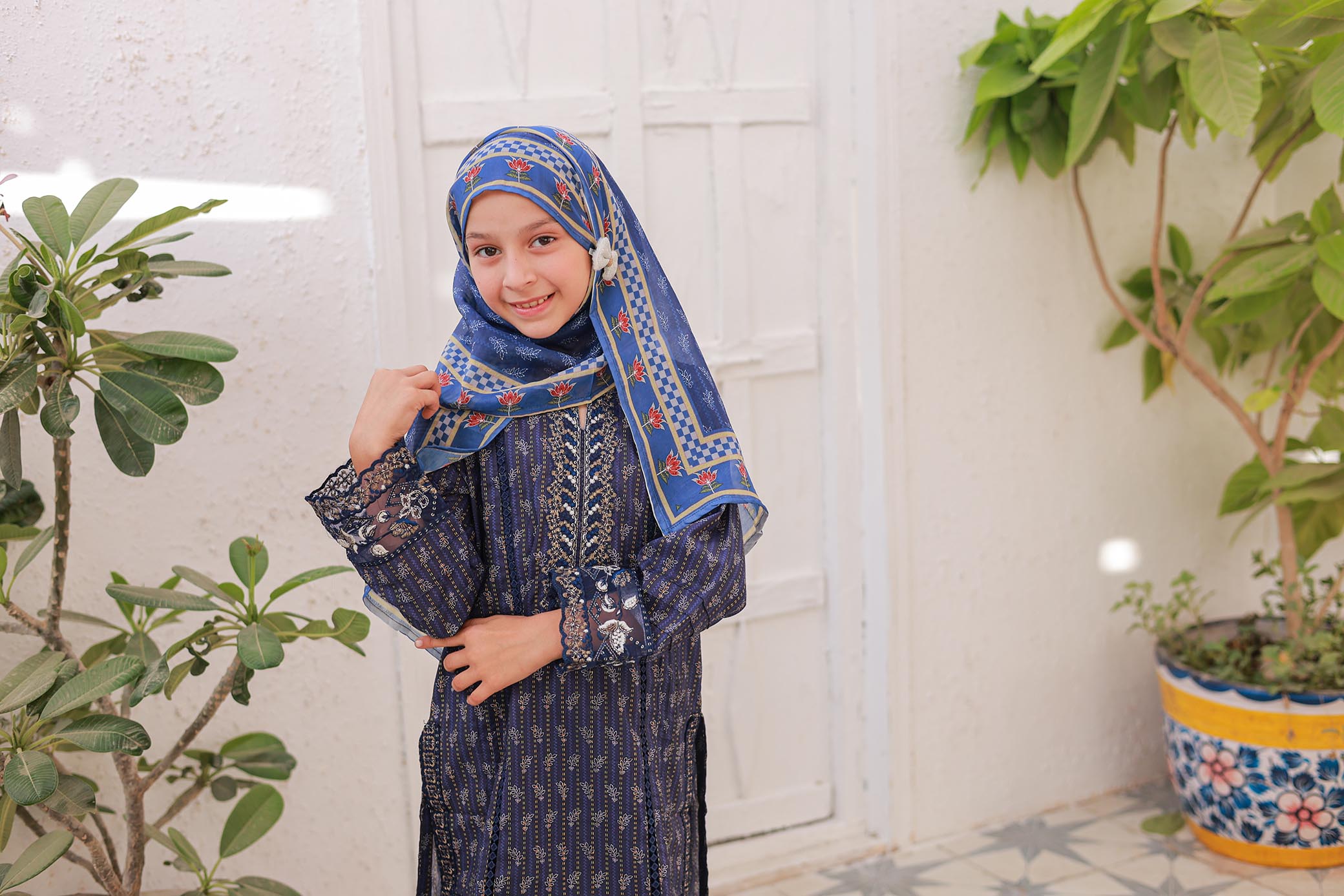 Child wearing a blue and white patterned hijab and dress, standing indoors with plants in the background.