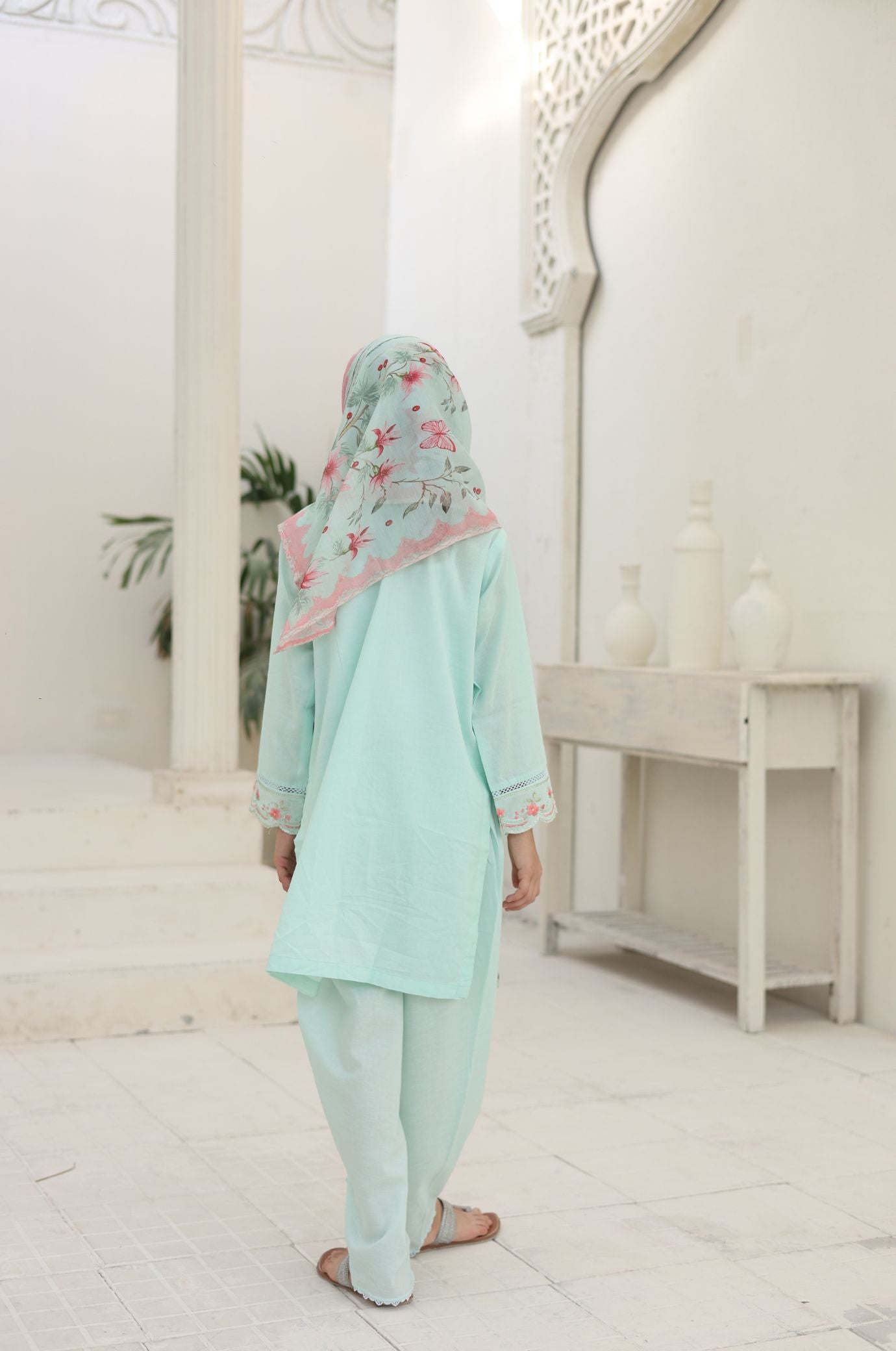 Person wearing a light blue traditional outfit with a floral headscarf in a white room.