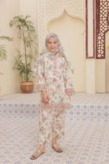 Young girl in a floral outfit standing in a decorated room with plants and architectural elements.