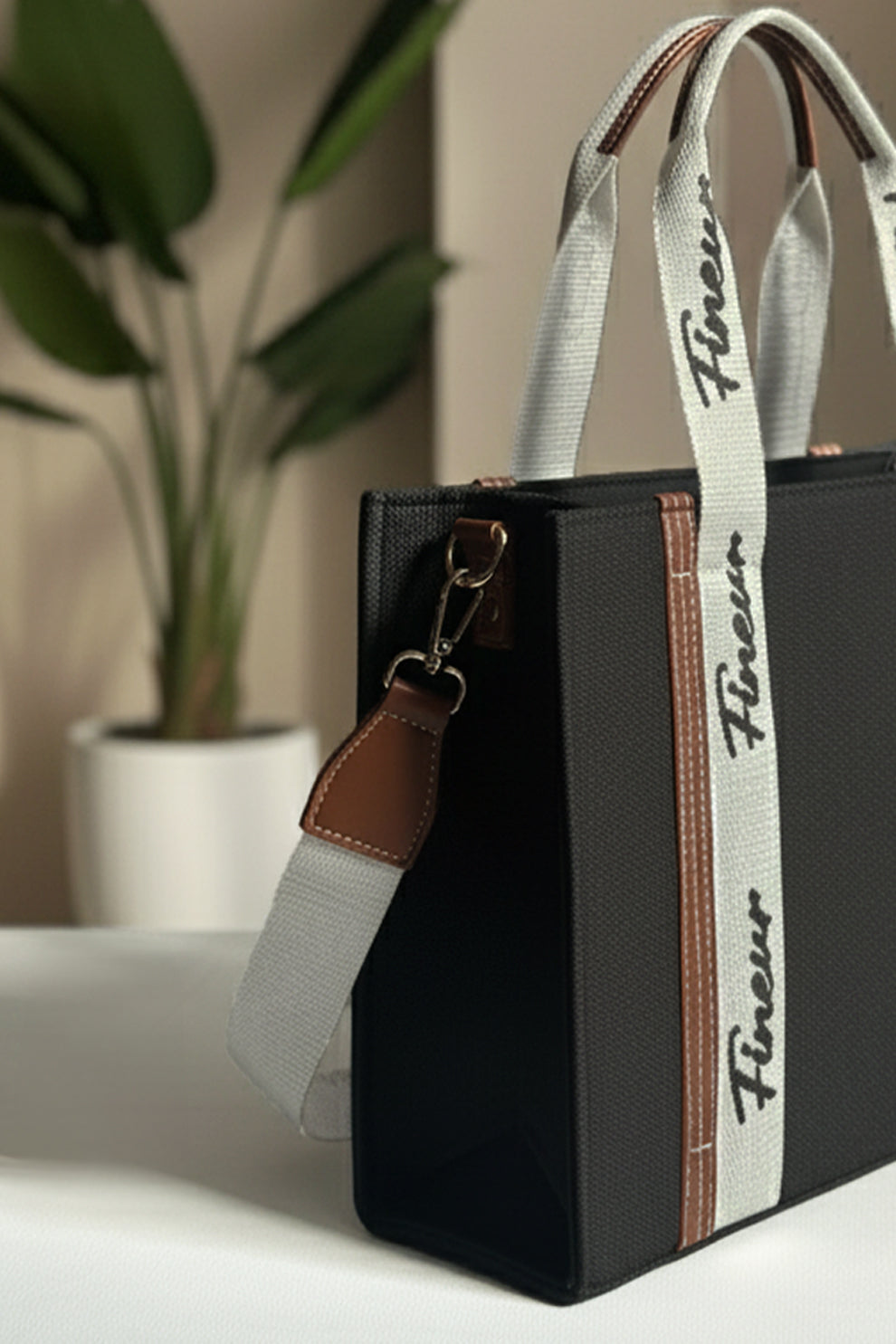 Black handbag with a white strap featuring 'fineur' branding, placed on a surface with a plant in the background.