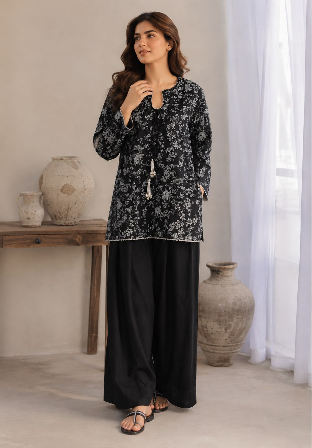 A woman models a black and gray floral printed long-sleeve tunic top paired with wide-leg black trousers, standing in a neutral-toned room with decorative vases and sheer curtains.