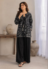 A woman models a black and gray floral printed long-sleeve tunic top paired with wide-leg black trousers, standing in a neutral-toned room with decorative vases and sheer curtains.