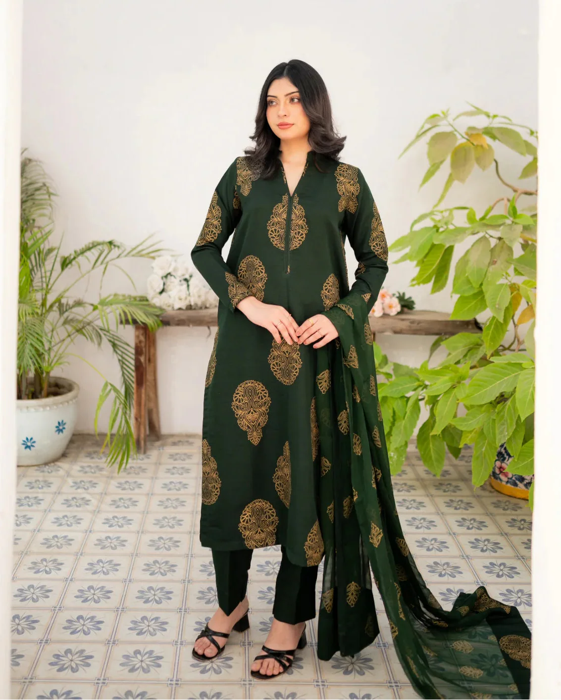 Woman wearing a bottle green traditional three piece suit with gold patterns in a room with plants.