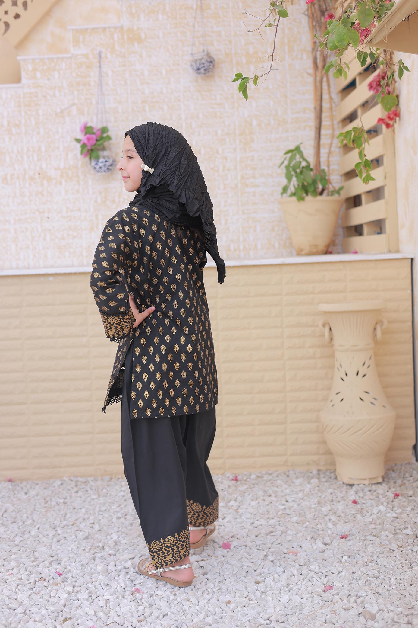 Woman in traditional outfit standing in a decorative outdoor setting with plants and a beige wall.