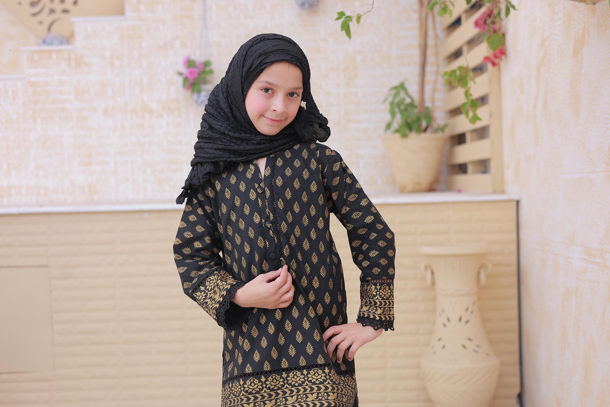 Young girl wearing a black and gold patterned outfit with a hijab in a room with decorative elements.