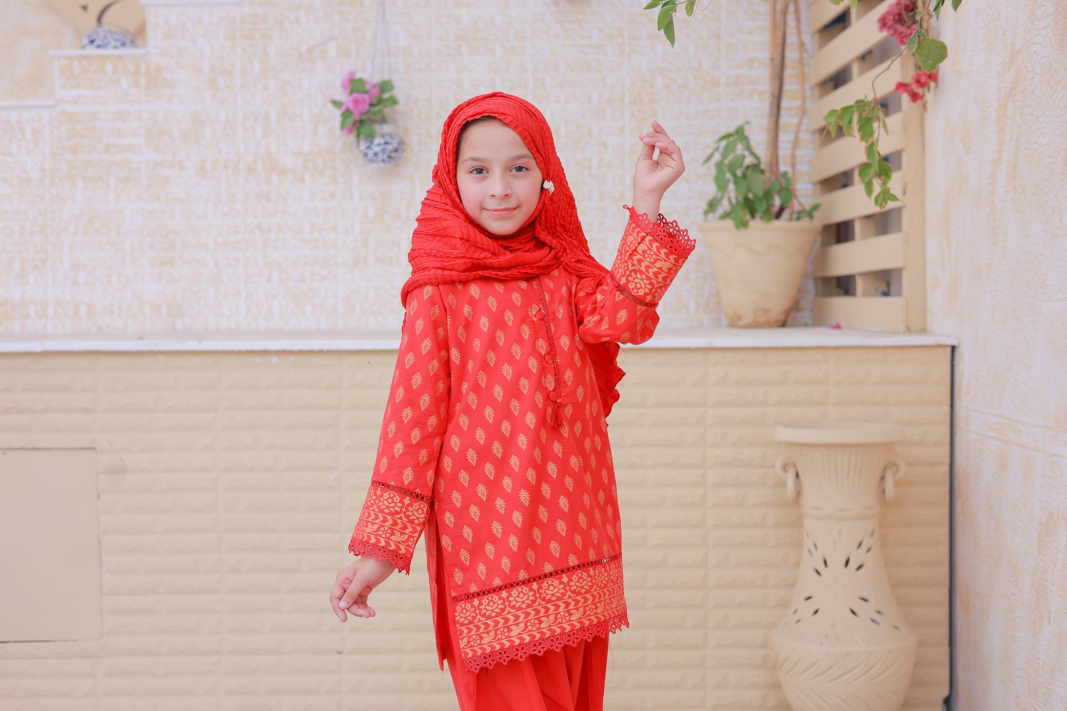 Child wearing a red traditional outfit with a matching headscarf against a light-colored wall.
