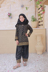 Young girl in a black and gold traditional outfit standing outdoors with decorative plants and a light-colored wall in the background.