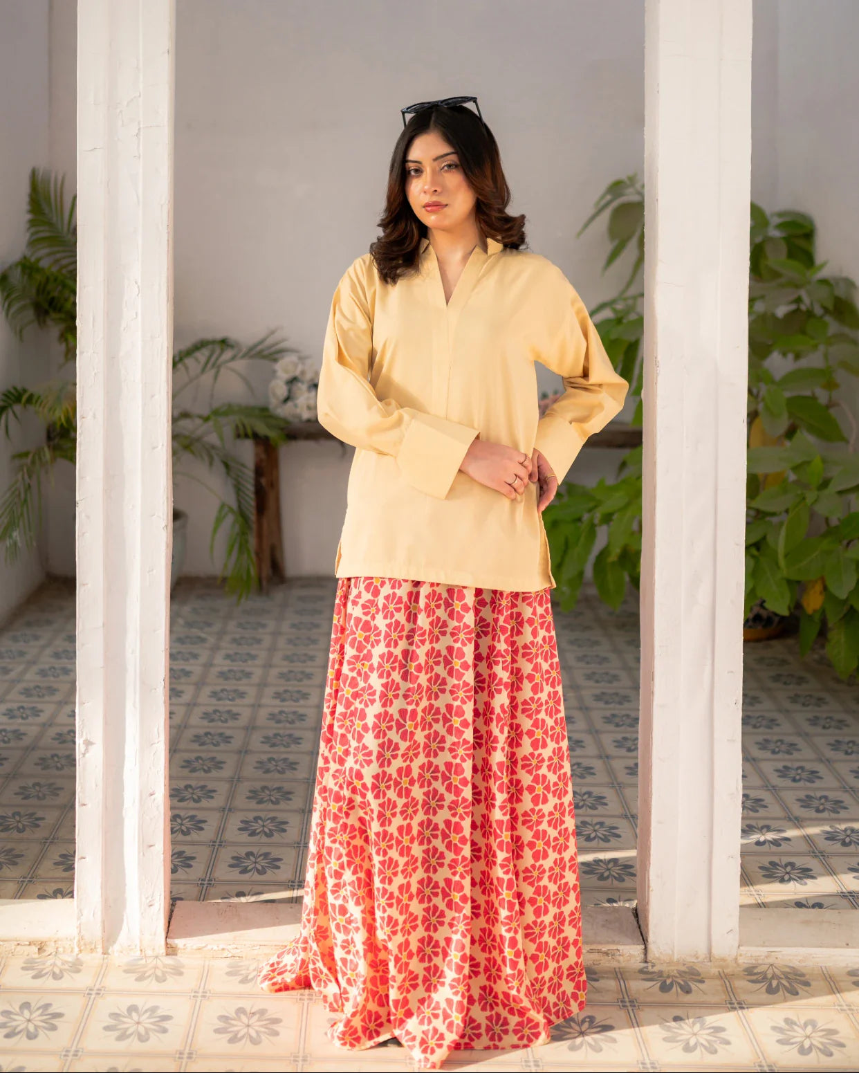 Woman wearing a lemon yellow top and red floral skirt standing in a room with plants.