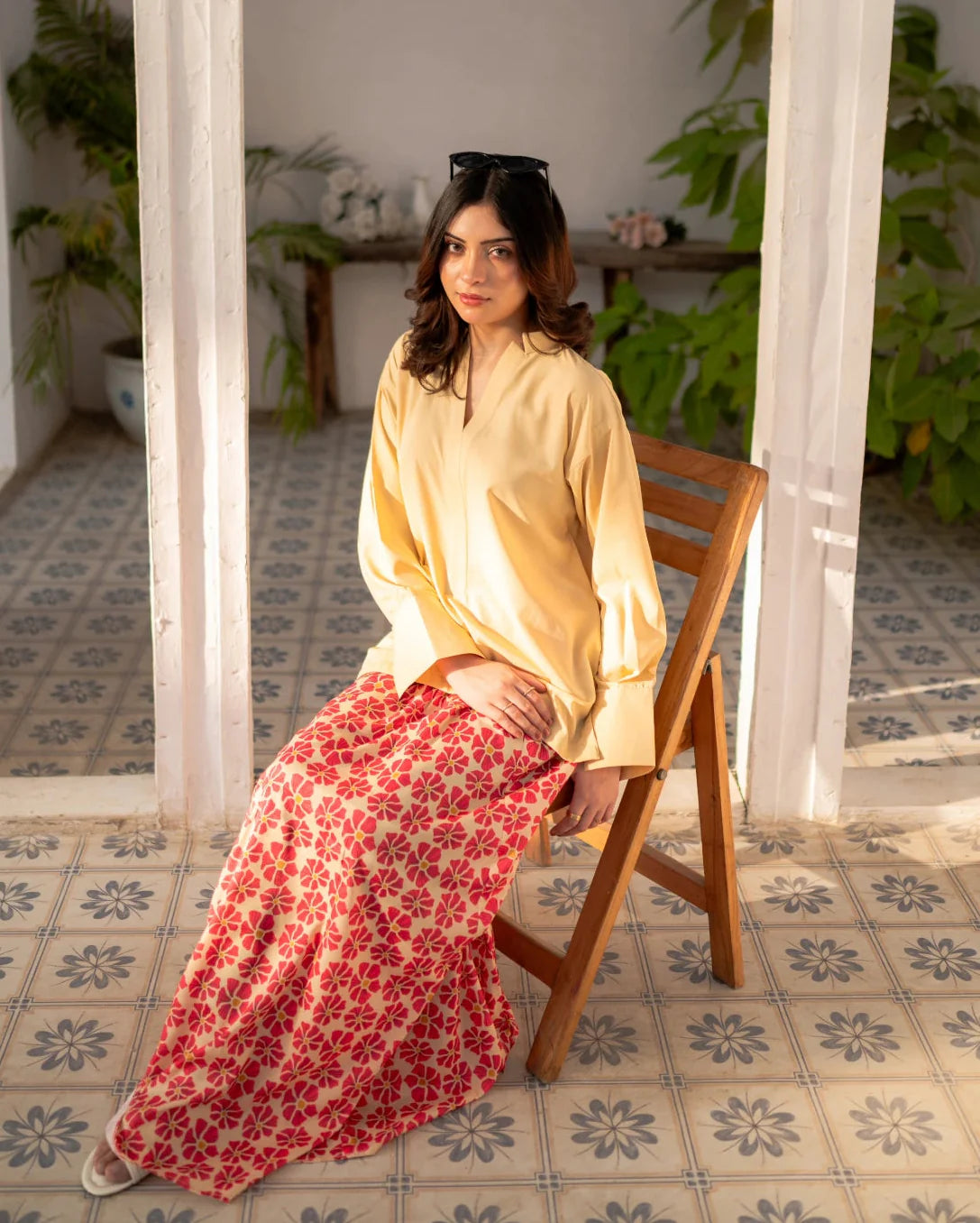Woman sitting on a wooden chair wearing a lemon yellow top and red floral skirt in a sunlit room with plants.