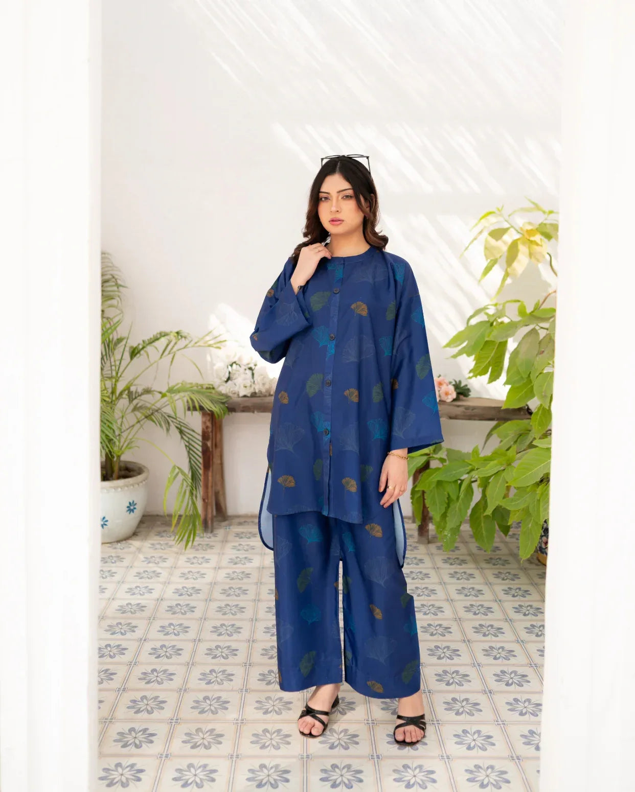Woman wearing a navy blue outfit with a pattern in a room with plants and a white wall.