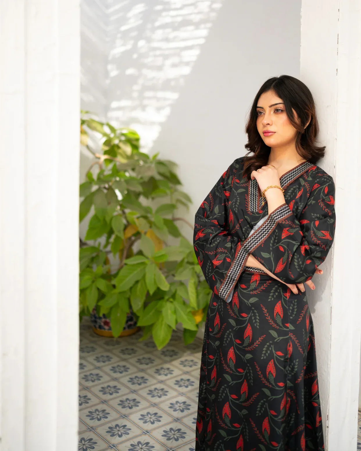 Woman in a black printed dress standing indoors with a plant and white wall in the background