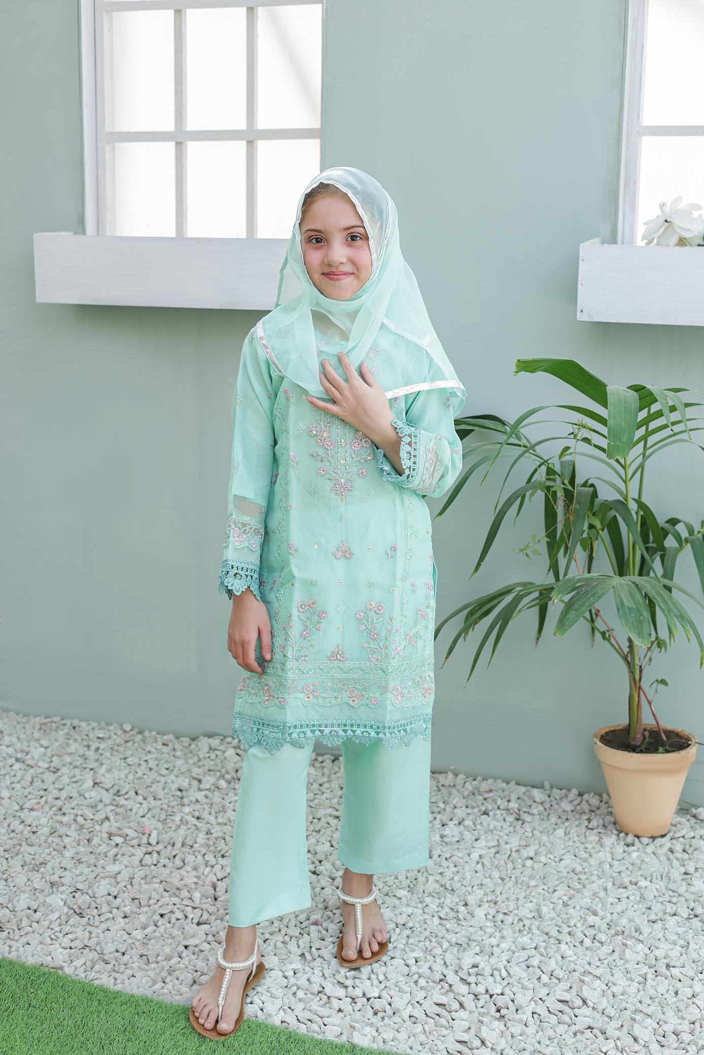Woman wearing a light green traditional outfit with a headscarf, standing in a room with a plant.