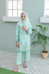 Woman wearing a light green traditional outfit with a headscarf, standing in a room with a plant.