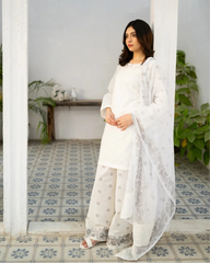 Woman in a white kurta set standing in a room with decorative tiles and plants.
