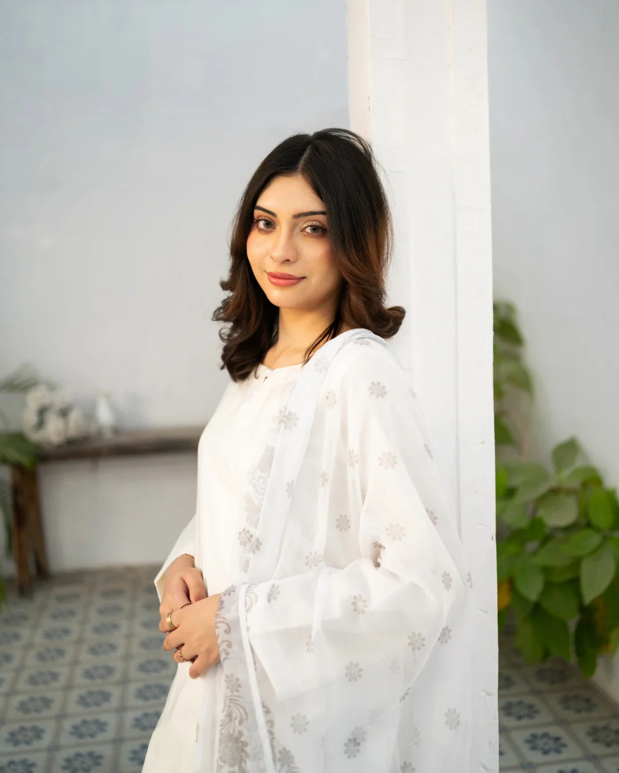 Woman in a white kurta set standing against a light-colored wall with plants in the background.
