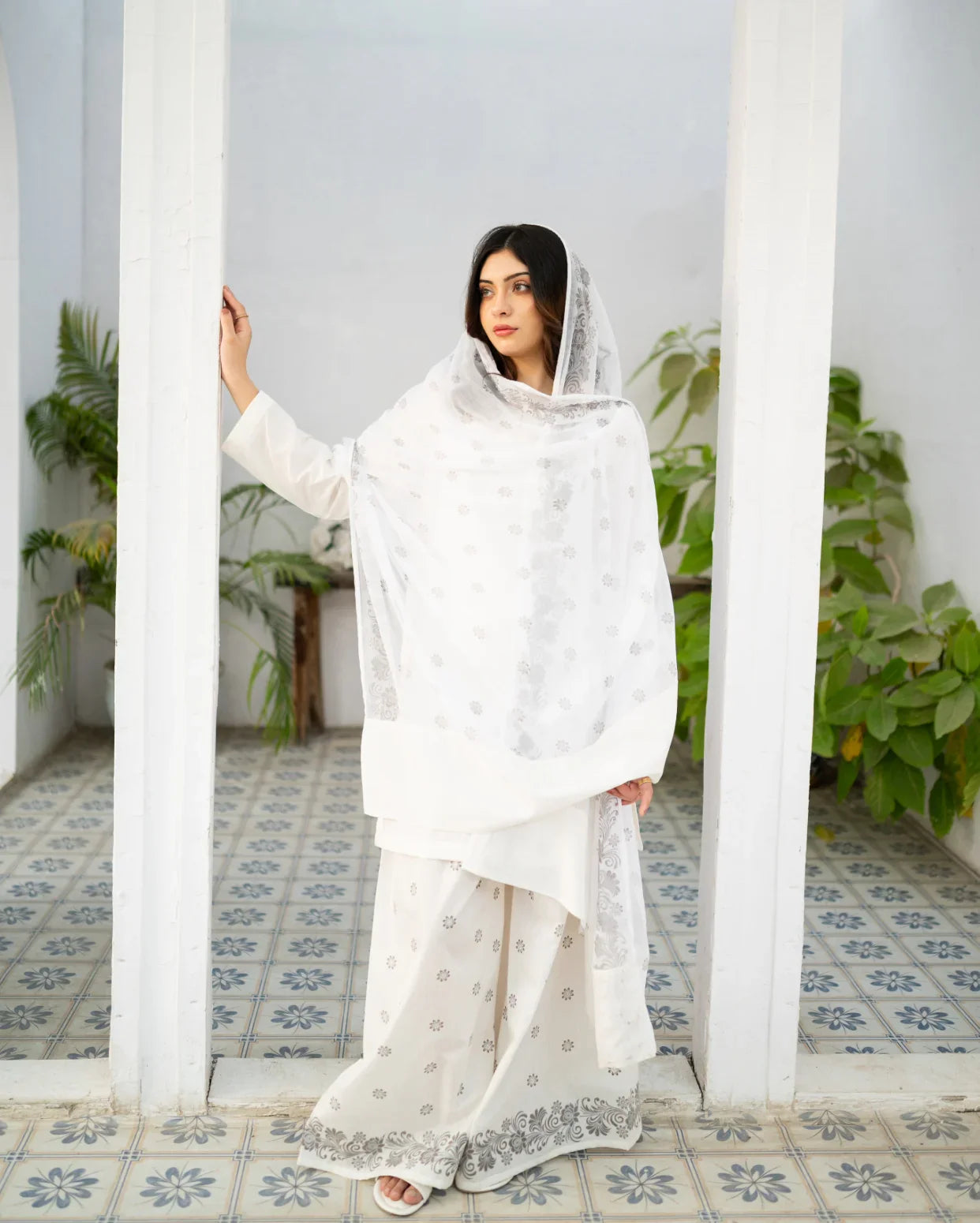Woman in a white traditional kurta set standing in a room with plants and patterned floor.