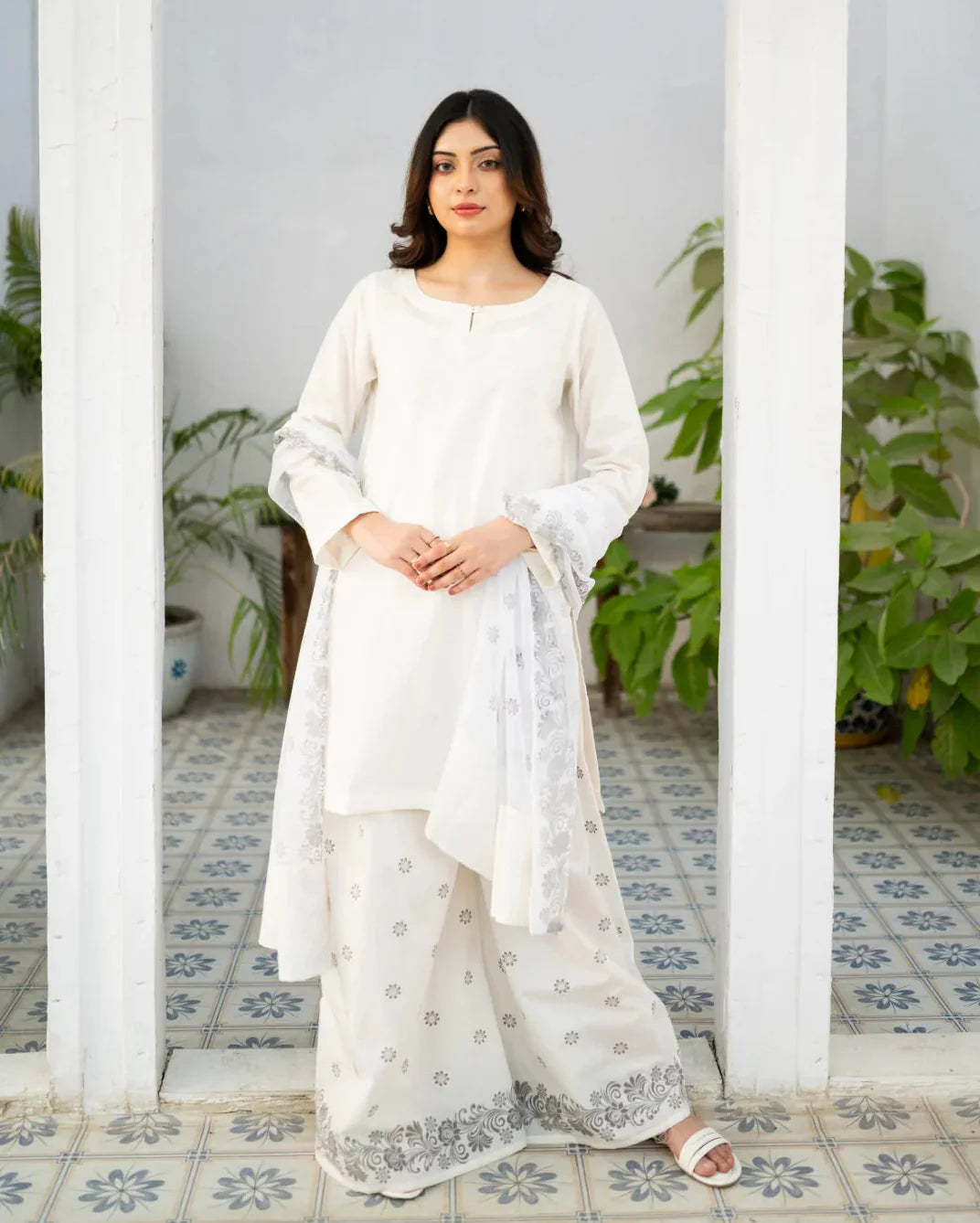 Woman in a white traditional kurta set standing in a room with plants and patterned floor.